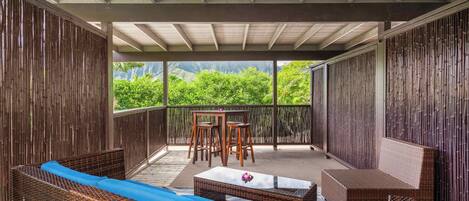 A covered outdoor seating area with wicker furniture and a table, overlooking lush greenery.