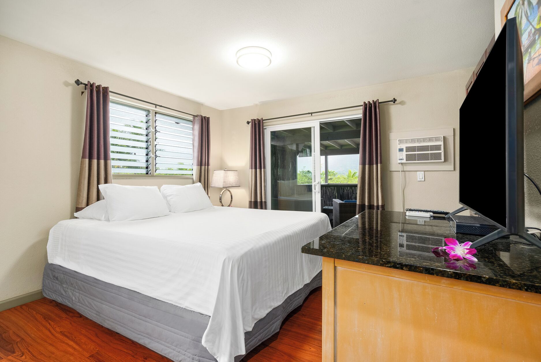 A hotel room with a bed, a desk with a flat-screen TV, a window with curtains, and an air conditioning unit.