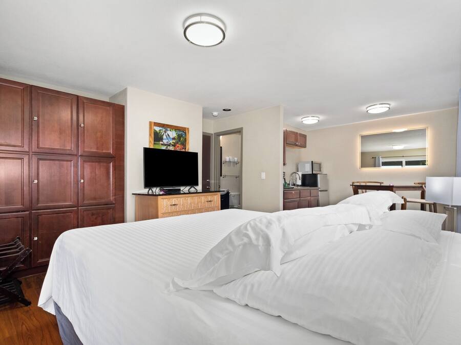 A hotel room with a large bed, wooden wardrobe, a flat-screen TV, a small kitchenette, and a bathroom with a mirror.