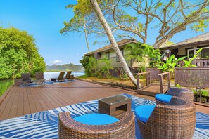 A wooden deck with wicker furniture and a glass table, overlooking a body of water and a mountain range. - Paradise Bay Resort (Kaneohe)