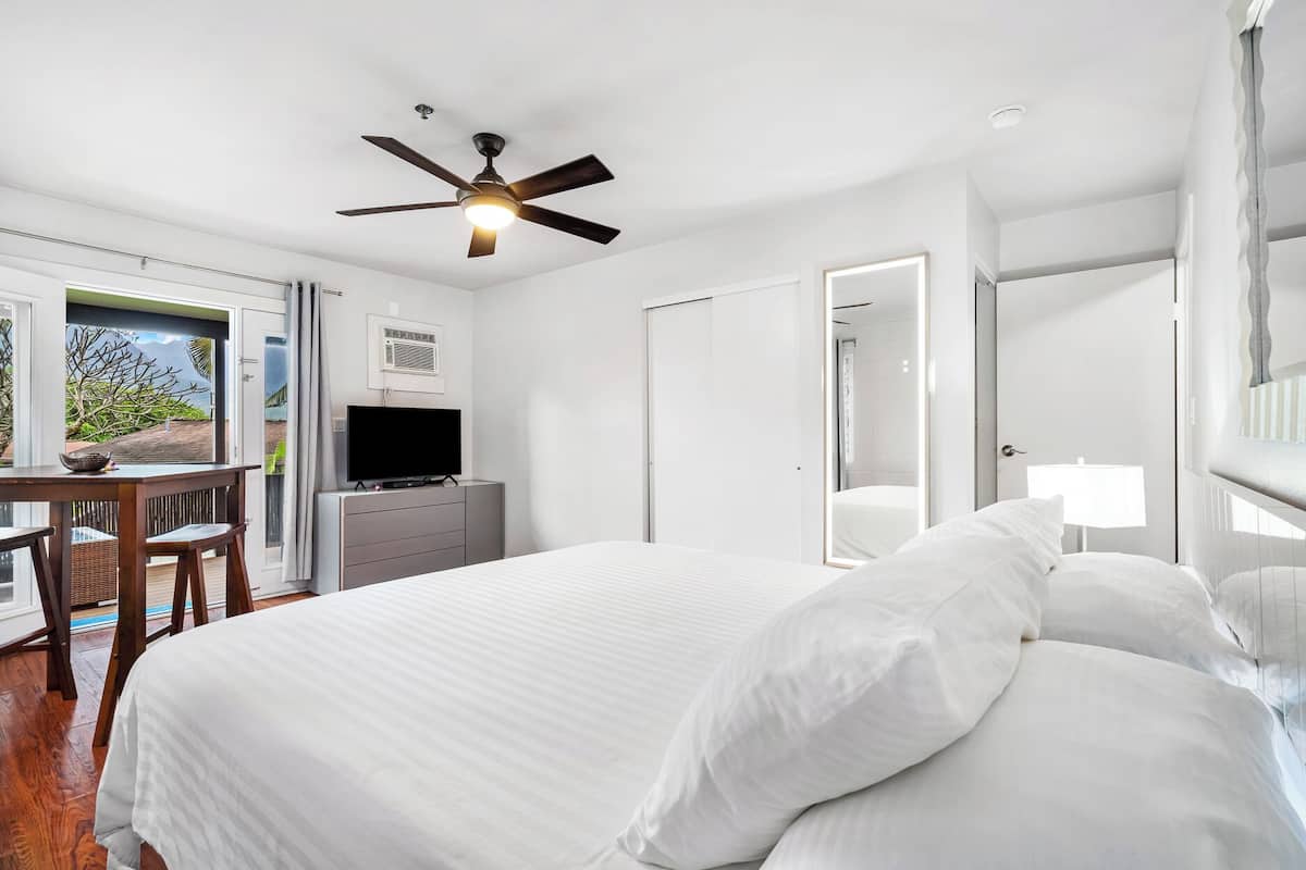 A bedroom with a bed, a desk with a television, a ceiling fan, and a view of the outdoors.