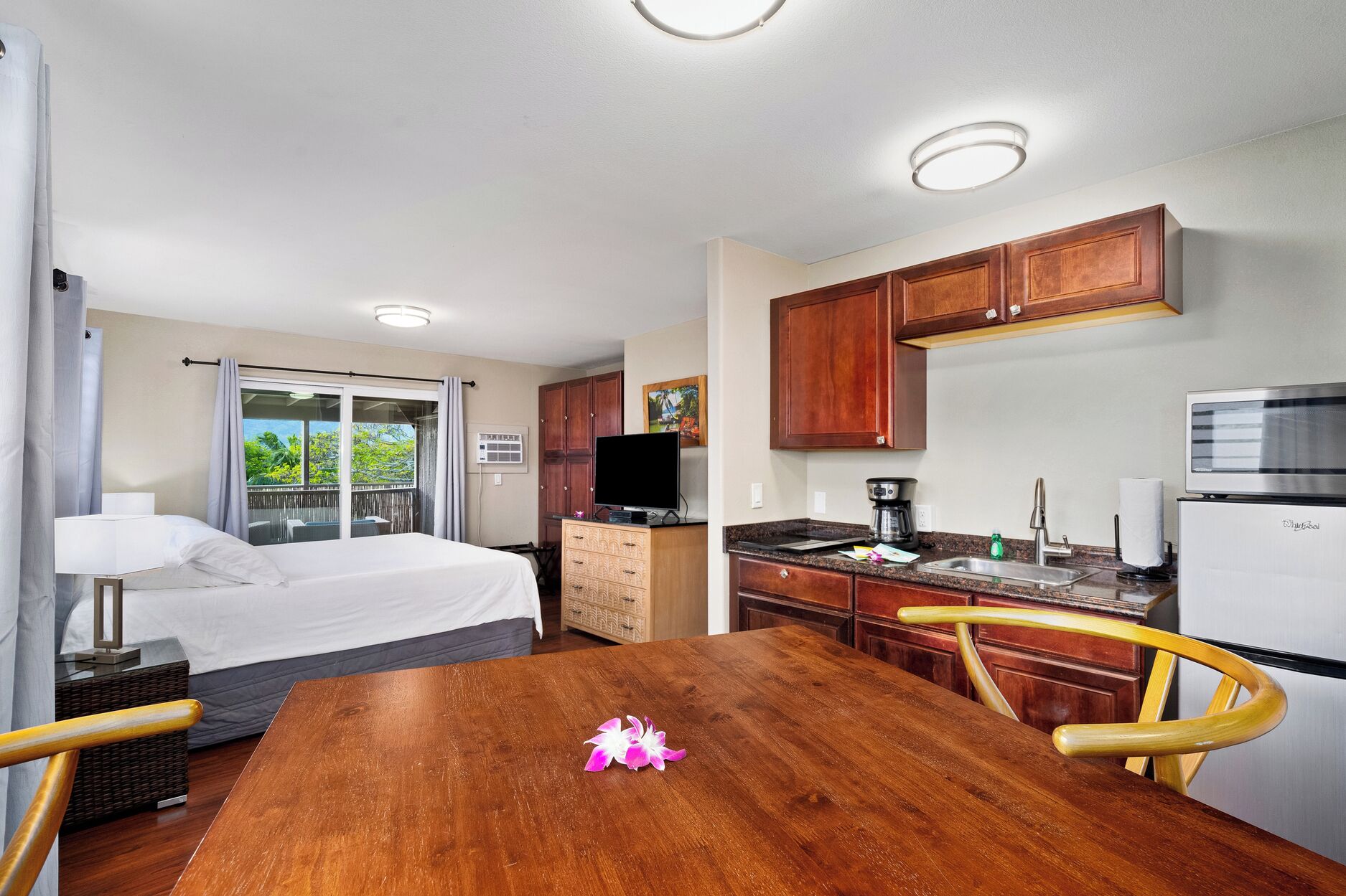 A hotel room with a bed, a wooden dining table, a kitchen area with wooden cabinets, a refrigerator, and a television.