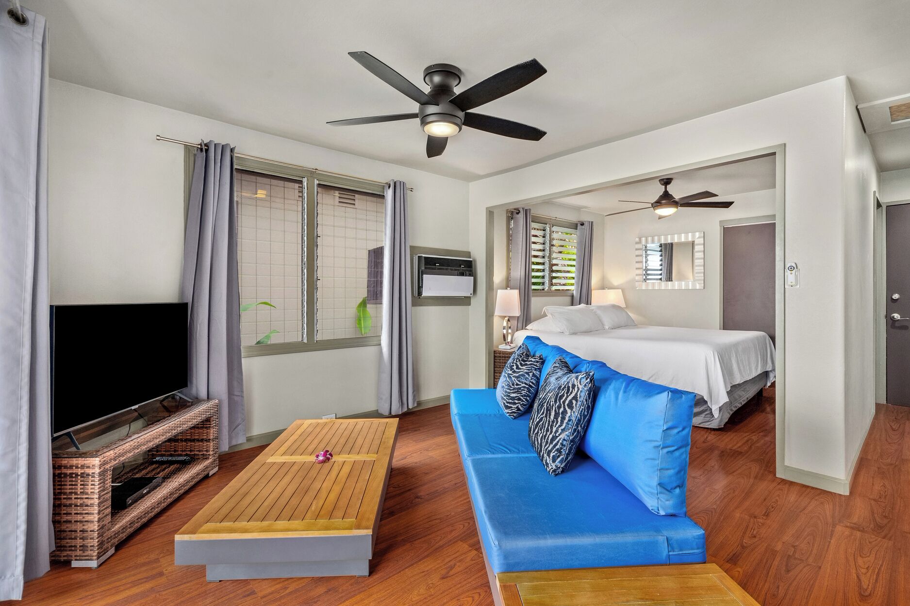 A modern hotel room with a blue sofa, a wooden coffee table, a TV on a built-in shelf, two beds, ceiling fans, and a window with blinds.