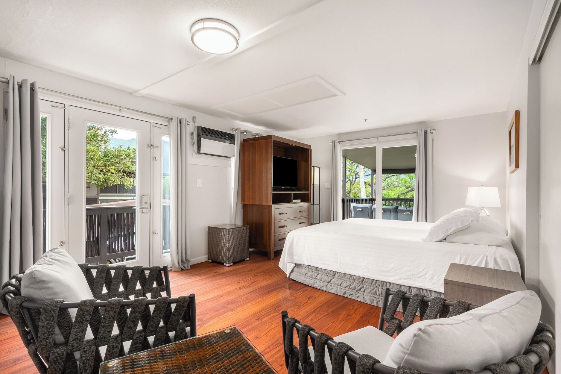 A hotel room with a bed, a TV, a seating area with chairs, and a balcony with a view of greenery.