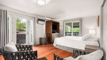 A hotel room with a bed, a TV, a seating area with chairs, and a balcony with a view of greenery.