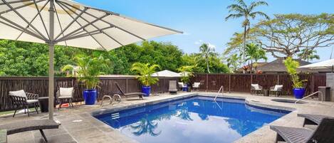 A swimming pool with lounge chairs and umbrellas, surrounded by a wooden fence and tropical plants.