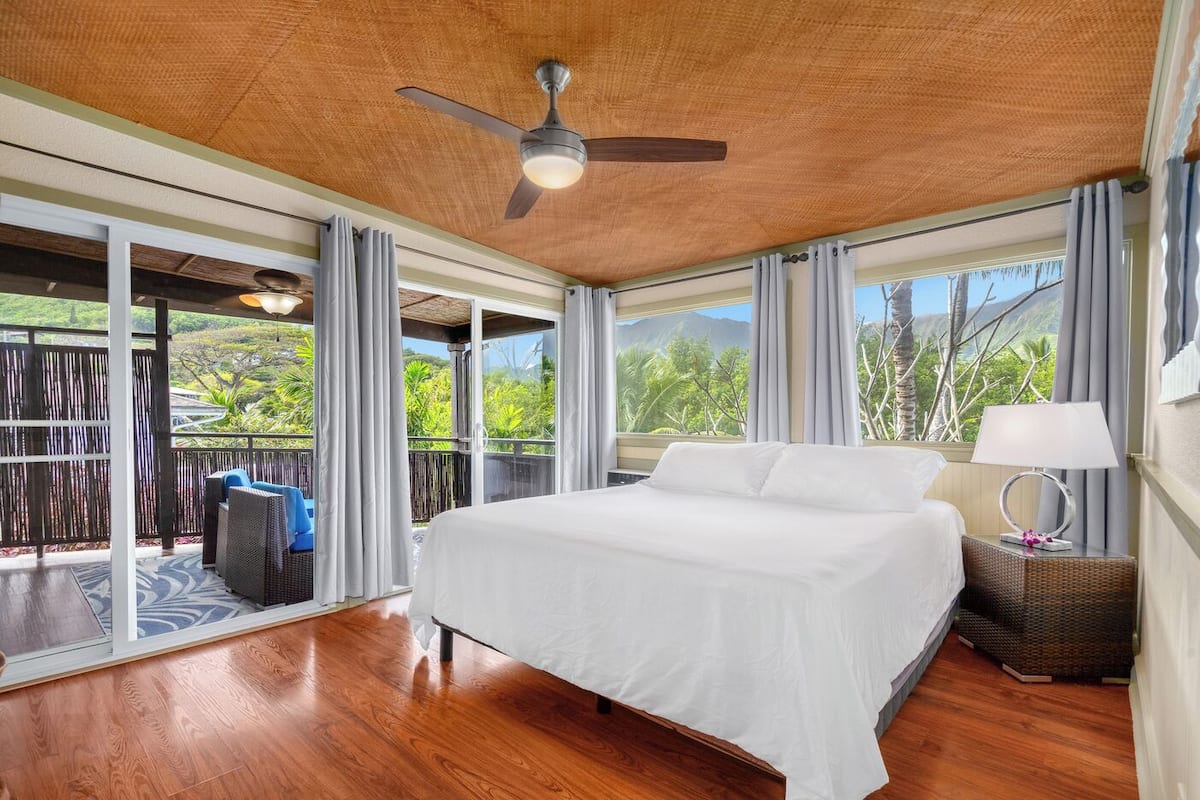 A bedroom with a large bed, a ceiling fan, and a view of lush greenery through large windows.