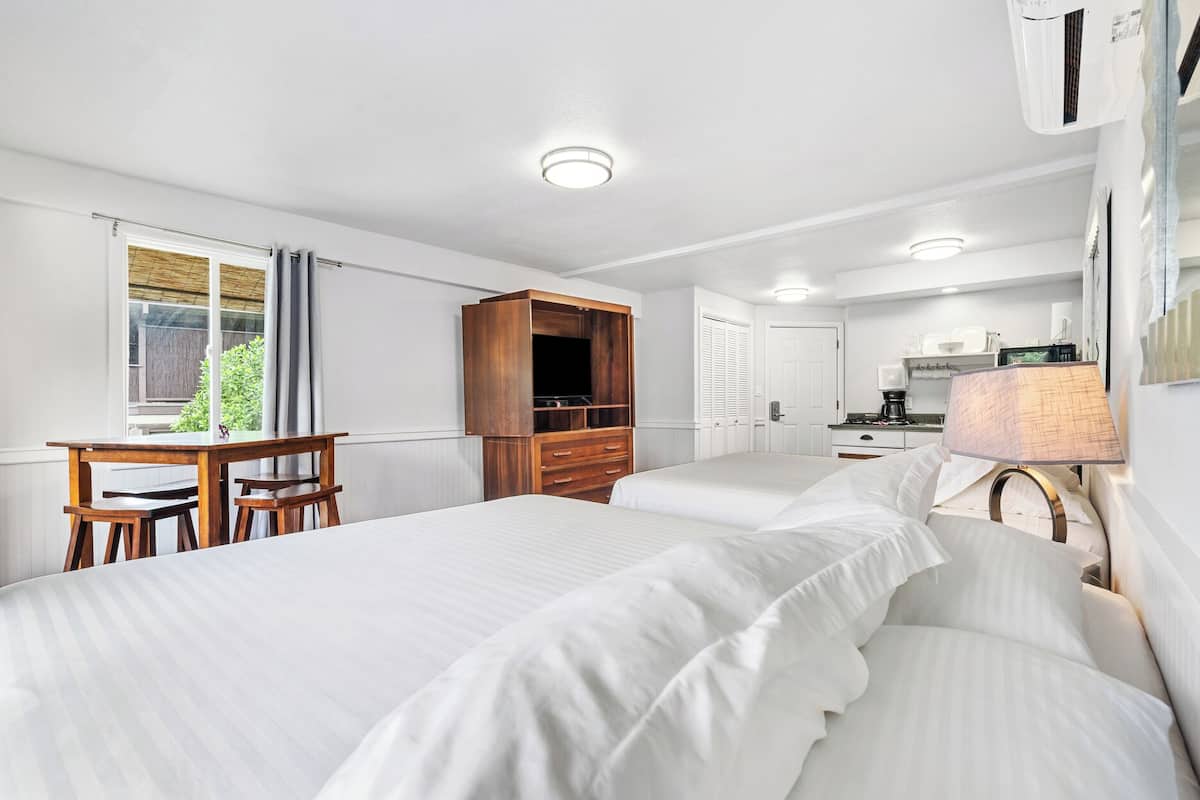 A hotel room with two beds, a wooden desk, a television, a kitchenette, and a window with curtains.