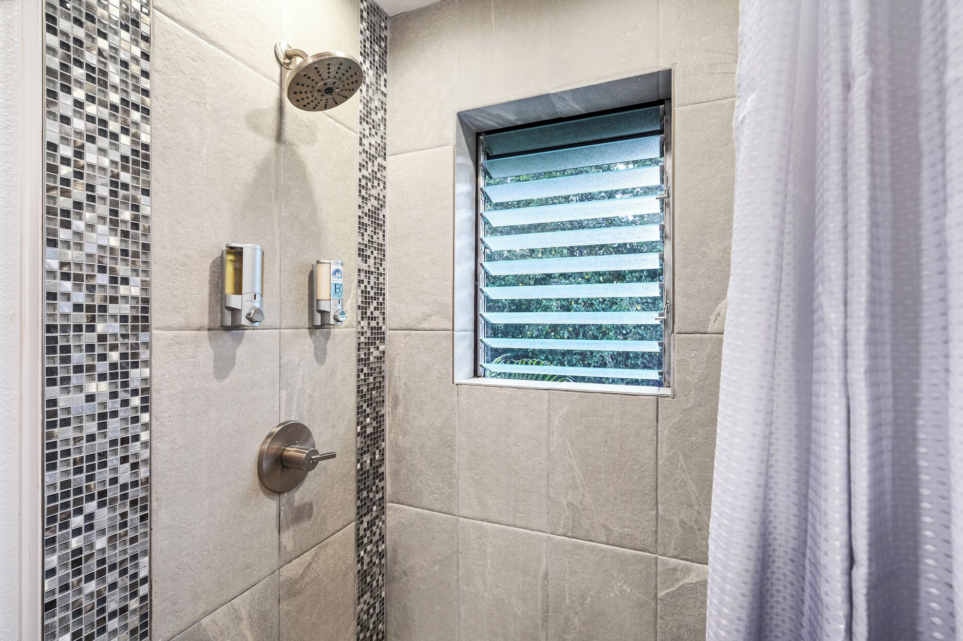Um chuveiro com um chuveiro de chuva, uma janela com persianas horizontais e um dispensador de sabonete fixado na parede.