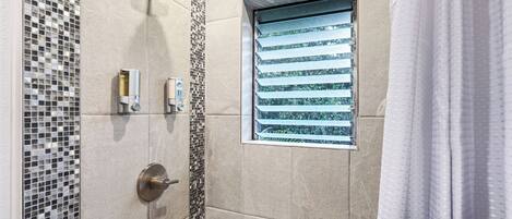 Een douche met regenstortdouche, een raam met horizontale zonwering en een zeepdispenser op de muur.