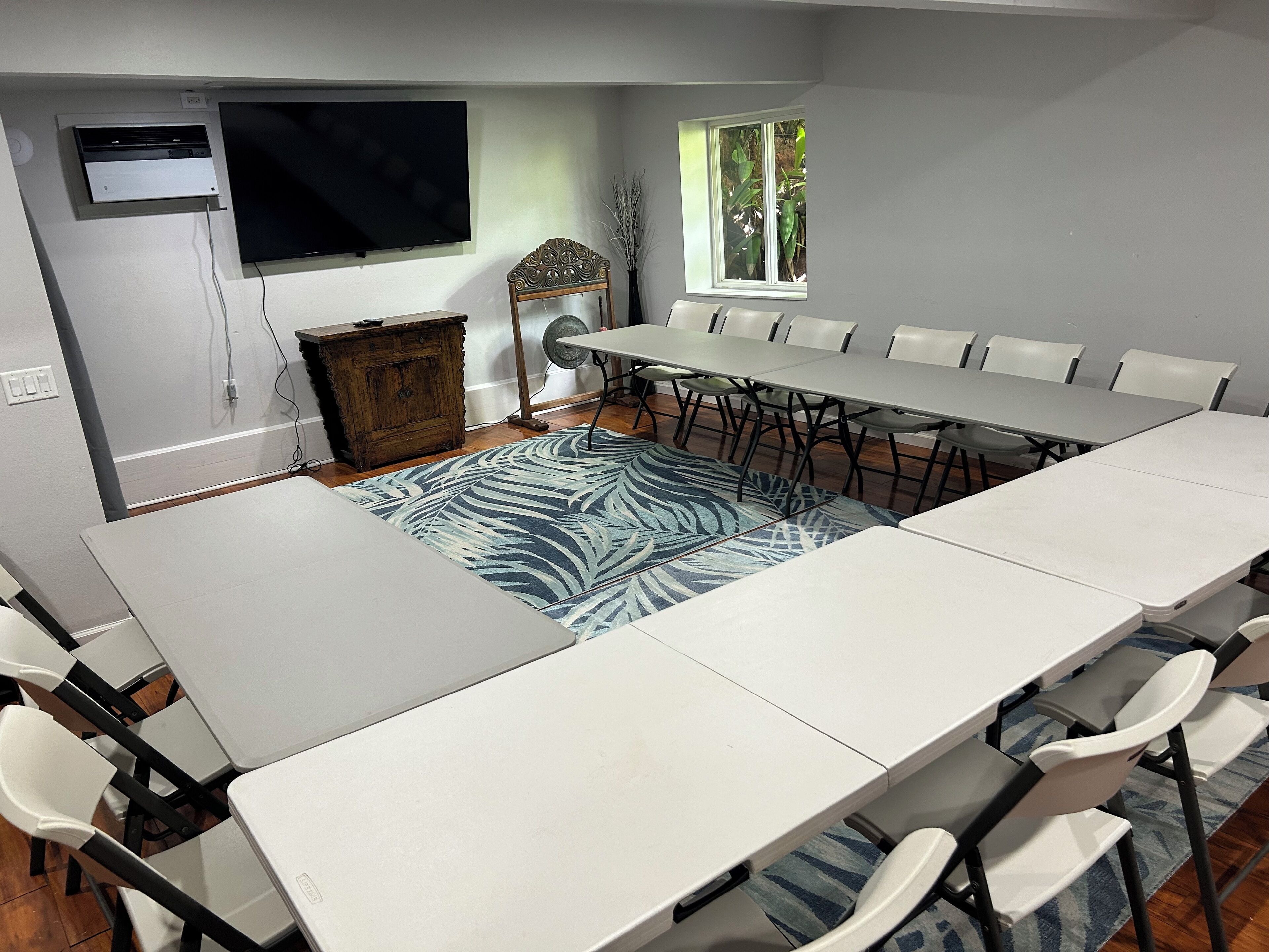 Un salón de conferencias con una larga mesa, sillas blancas, un televisor de pantalla plana, un armario de madera y una ventana con vistas a zonas verdes.