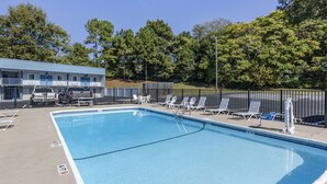 Una piscina rectangular con borde azul, rodeada por una valla negra. Hay sillones de descanso y una pequeña mesa con sillas a lo largo de la piscina.