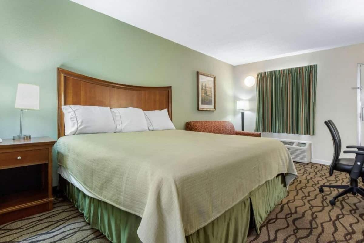 A hotel room with a large bed, a desk with a lamp, a chair, and a window with curtains.