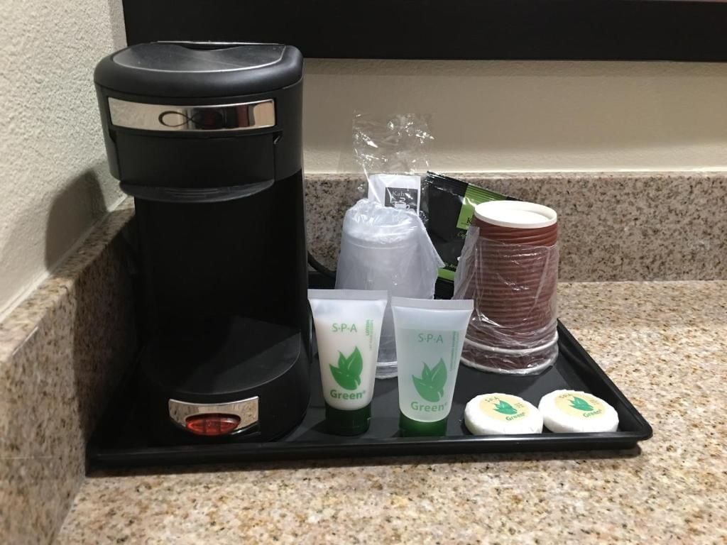 Una cafetera negra, dos productos de spa verdes y una pila de tazas rojas y blancas sobre una bandeja.