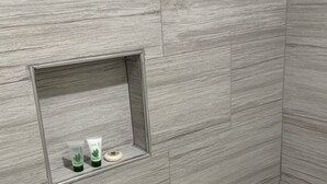 A bathroom with grey tiled walls and a white bathtub. There is a small shelf with a green bottle and a white tube. - Pacific Inn Anaheim (Anaheim)