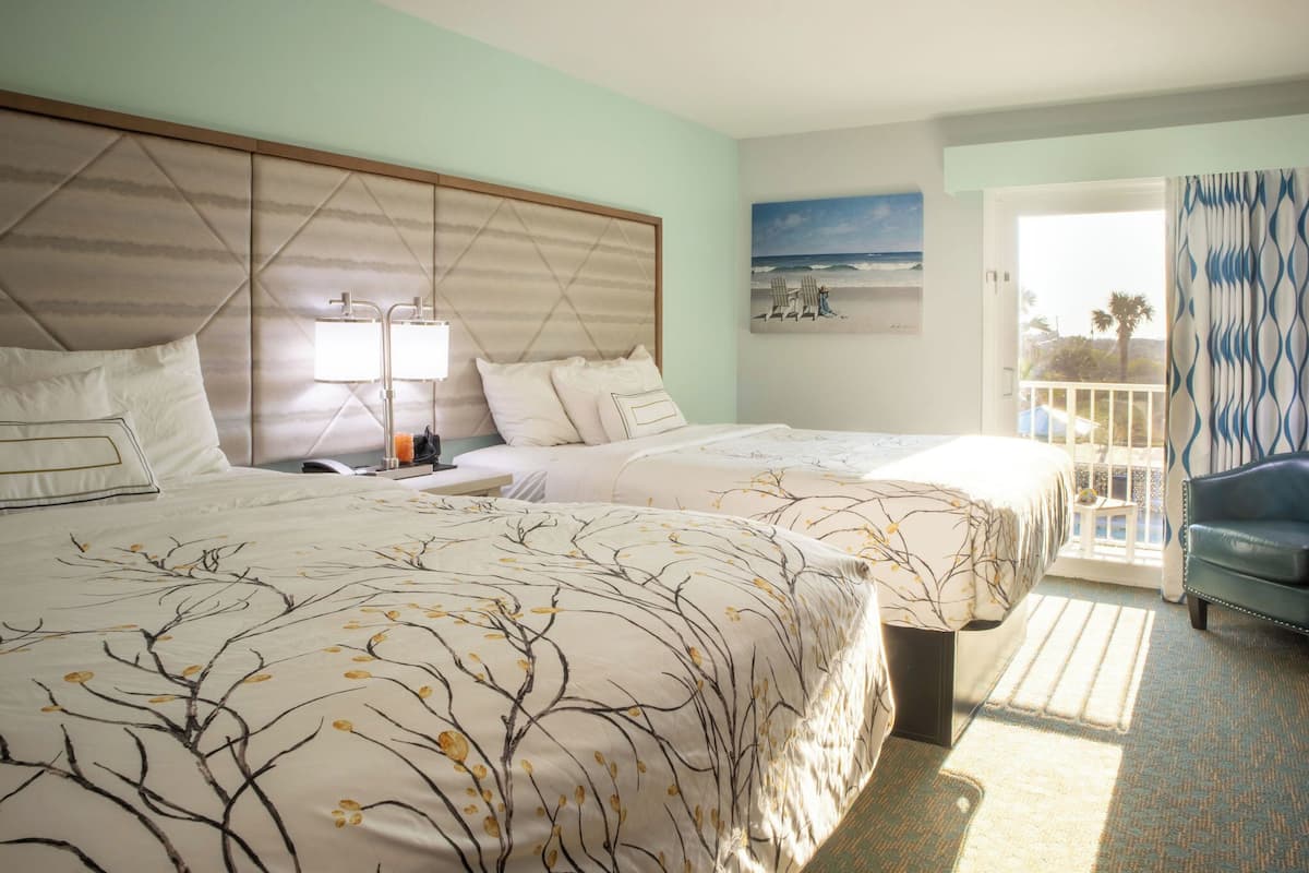 A hotel room with two beds, a nightstand, a lamp, a painting of a beach scene, and a view of the outdoors through a door.