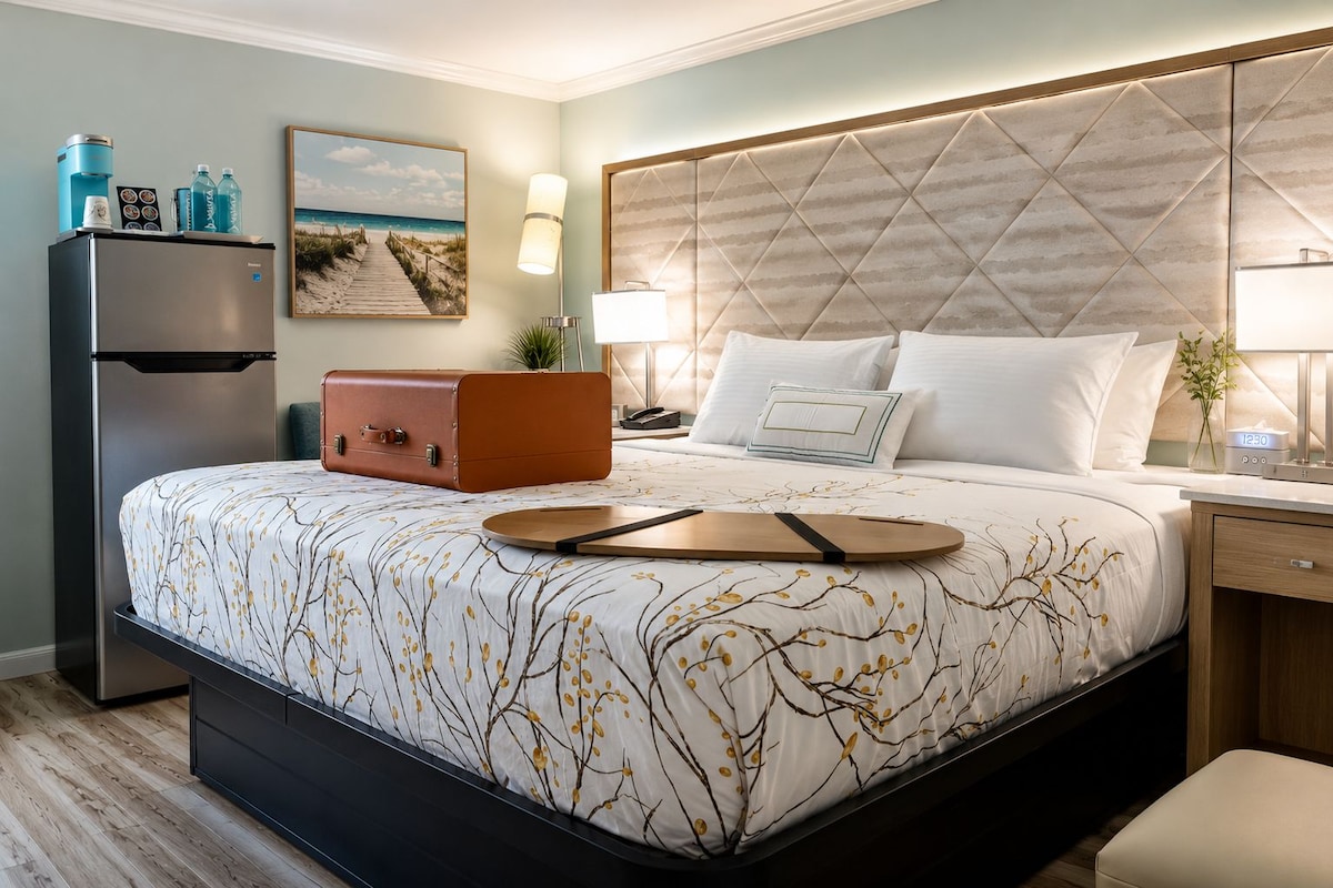 Ein Hotelzimmer mit Bett, Kühlschrank, Koffer, Lampe und einem Gemälde an der Wand.