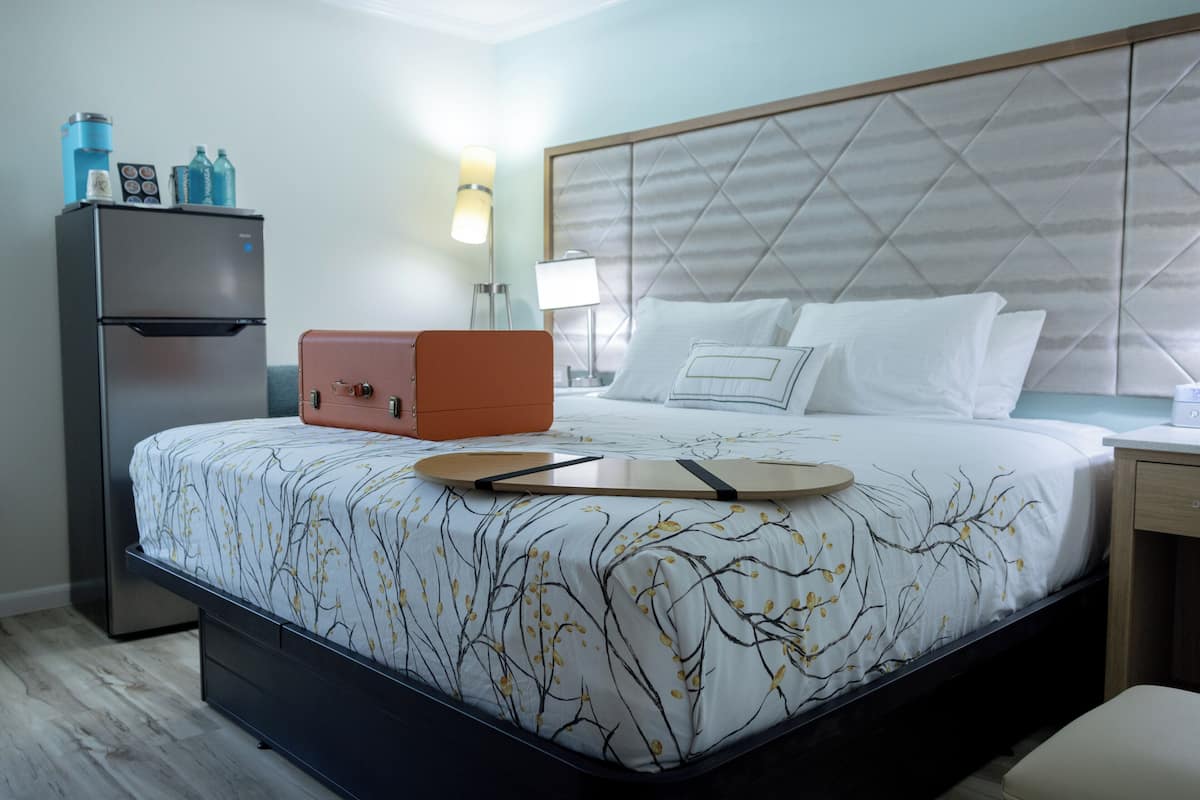 A hotel room with a bed, a nightstand, a lamp, a refrigerator, and a suitcase.