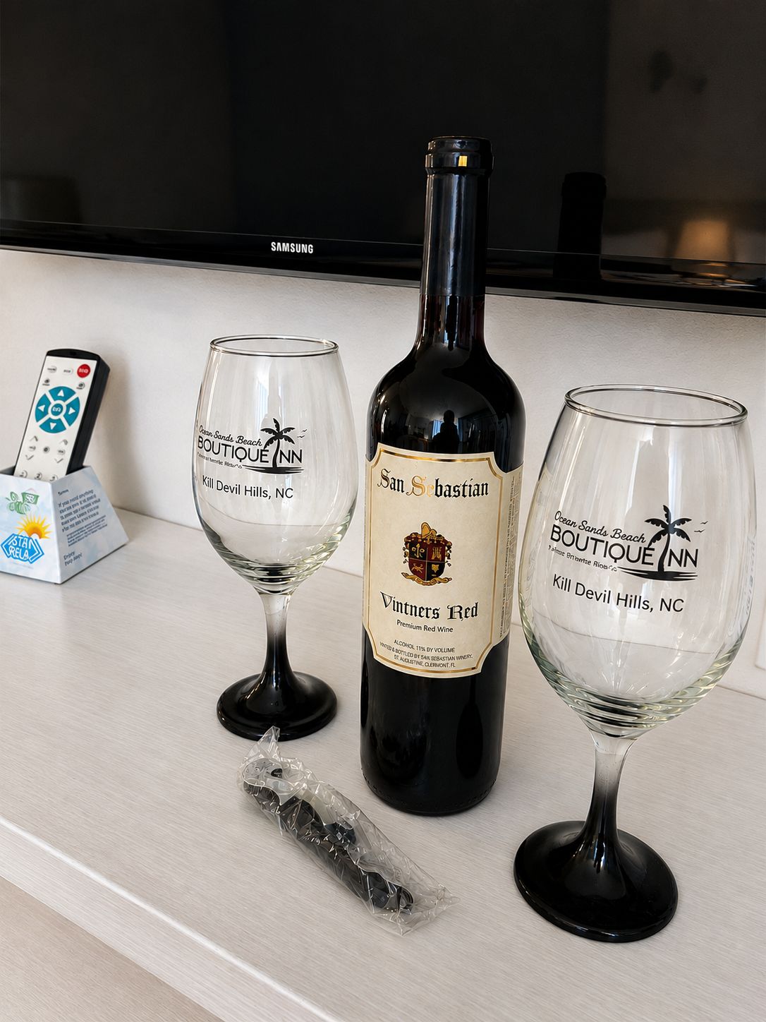 A bottle of Vintners Red wine, two glasses with the Boutique Inn logo, and a remote control on a counter.