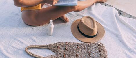 A person sitting on a towel by a pool, reading a book, with a hat and a netted bag nearby.