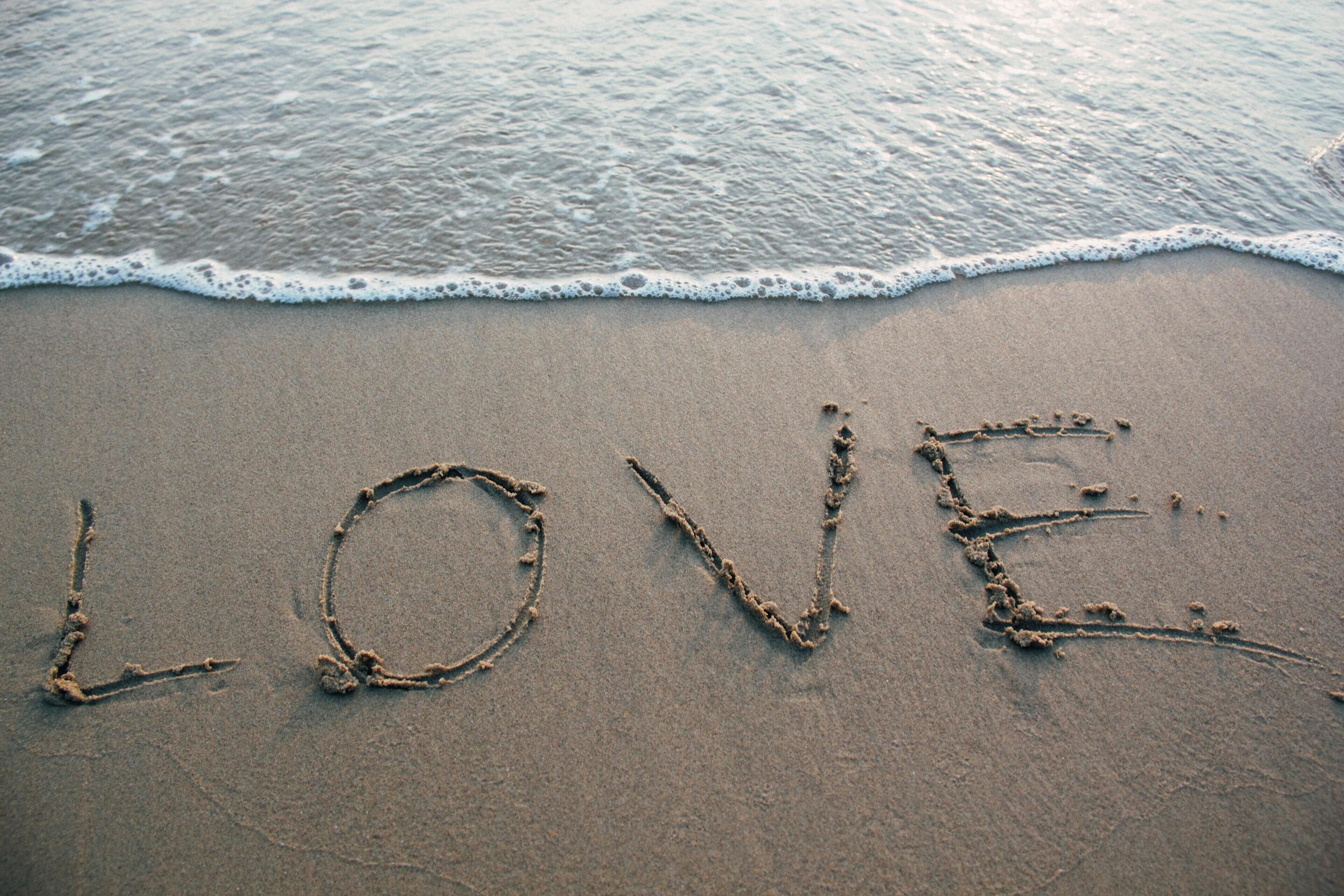The word "LOVE" written in the sand on a beach with waves in the background.