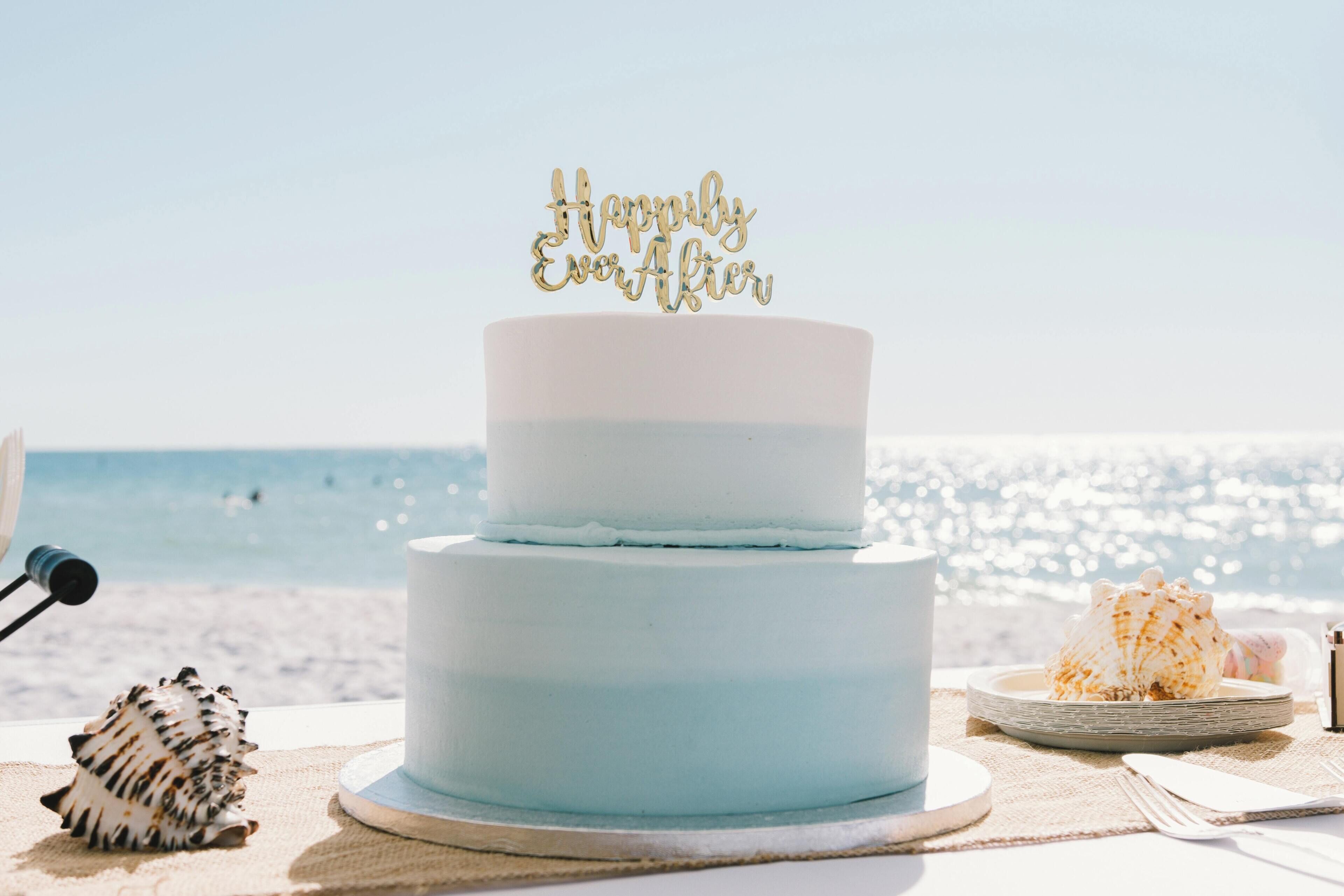 A two-tiered cake with a 'Happily Ever After' topper, placed on a table with a beach view in the background.
