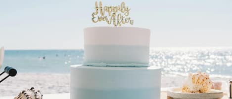 A two-tiered cake with a 'Happily Ever After' topper, placed on a table with a beach view in the background.