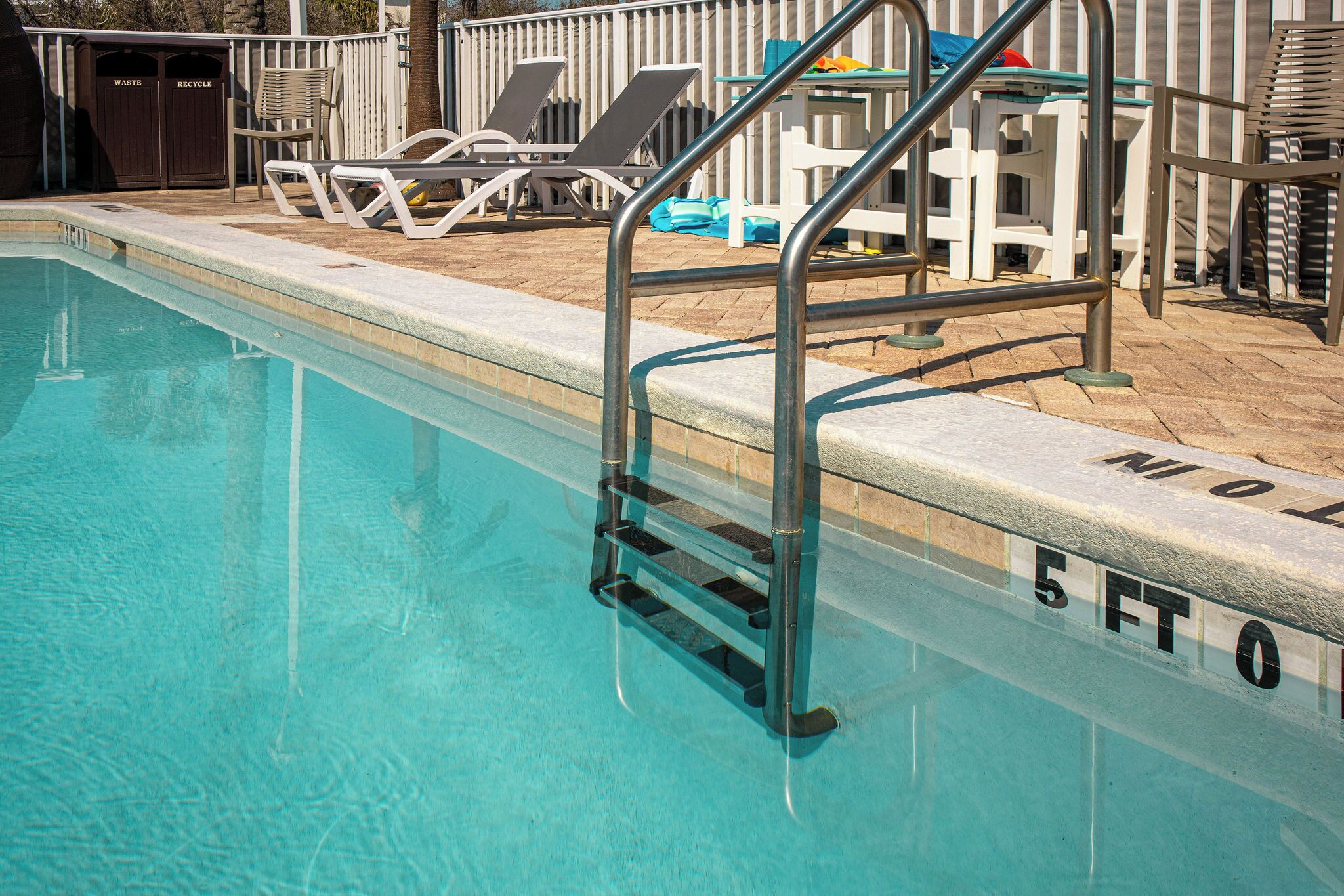 A swimming pool with a metal ladder and lounge chairs. There are trash bins labeled 'WASTE' and 'RECYCLE'.