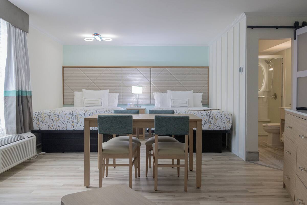 A modern hotel room with a large bed, a dining table with chairs, a bathroom with a shower, and a view of the city.