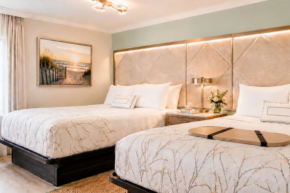 A hotel room with two beds, a nightstand with a lamp, a framed picture on the wall, and a tray on the bed.