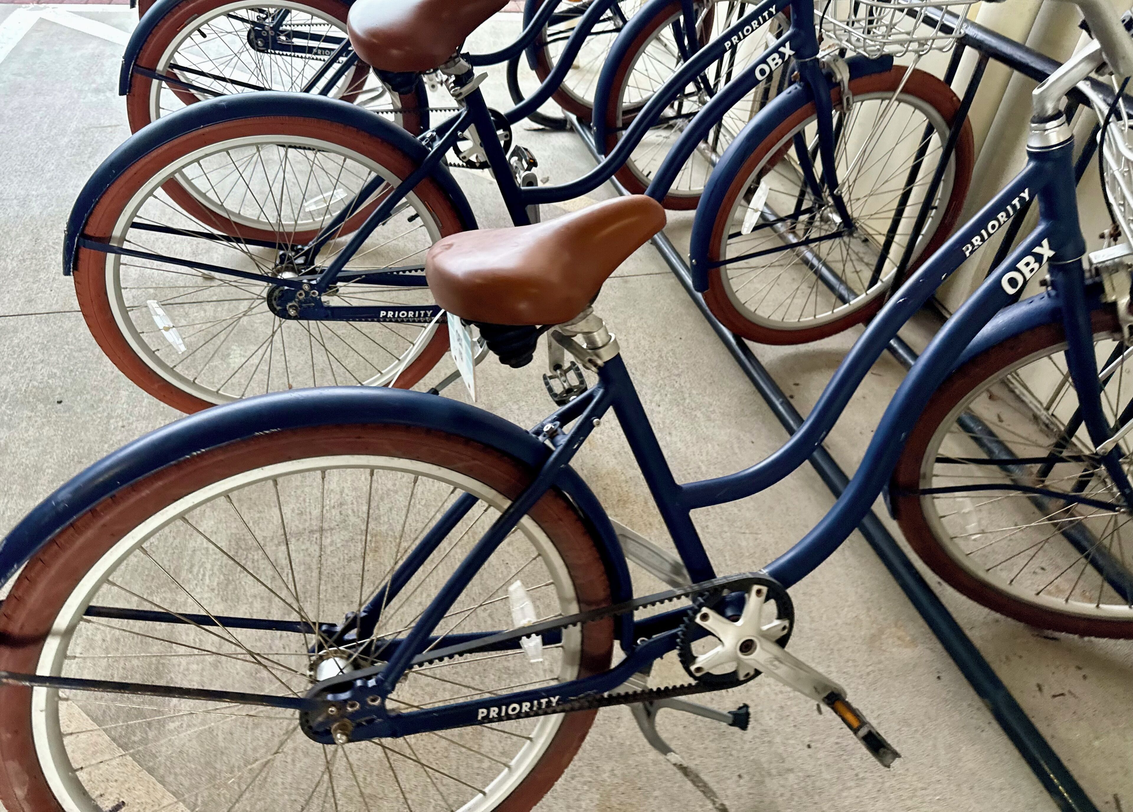 A blue bicycle with a brown seat and "PRIORITY" written on the frame, parked next to another bicycle.