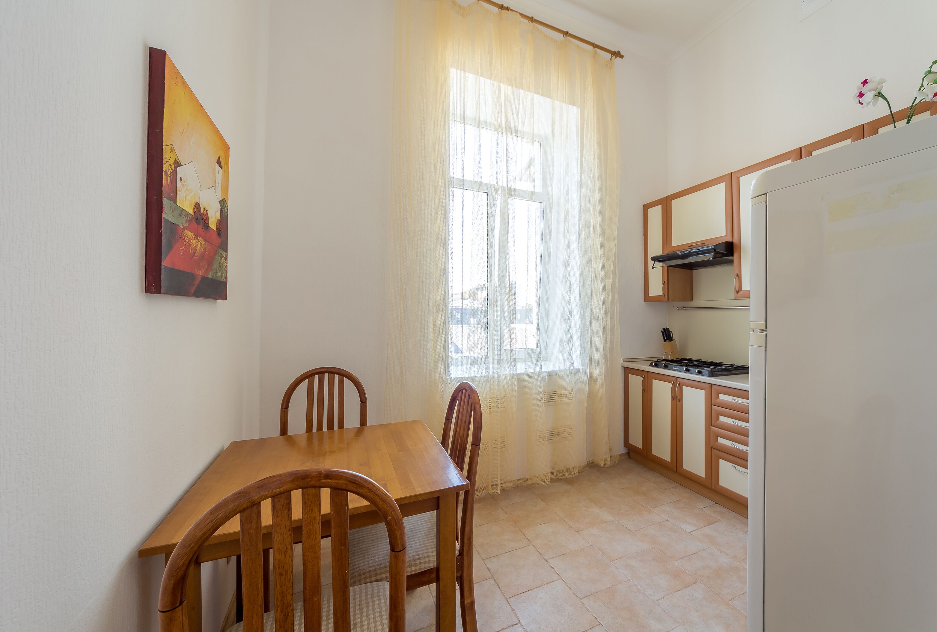 Une petite cuisine avec une table et des chaises, une fenêtre ornée de rideaux légers et un tableau accroché au mur.