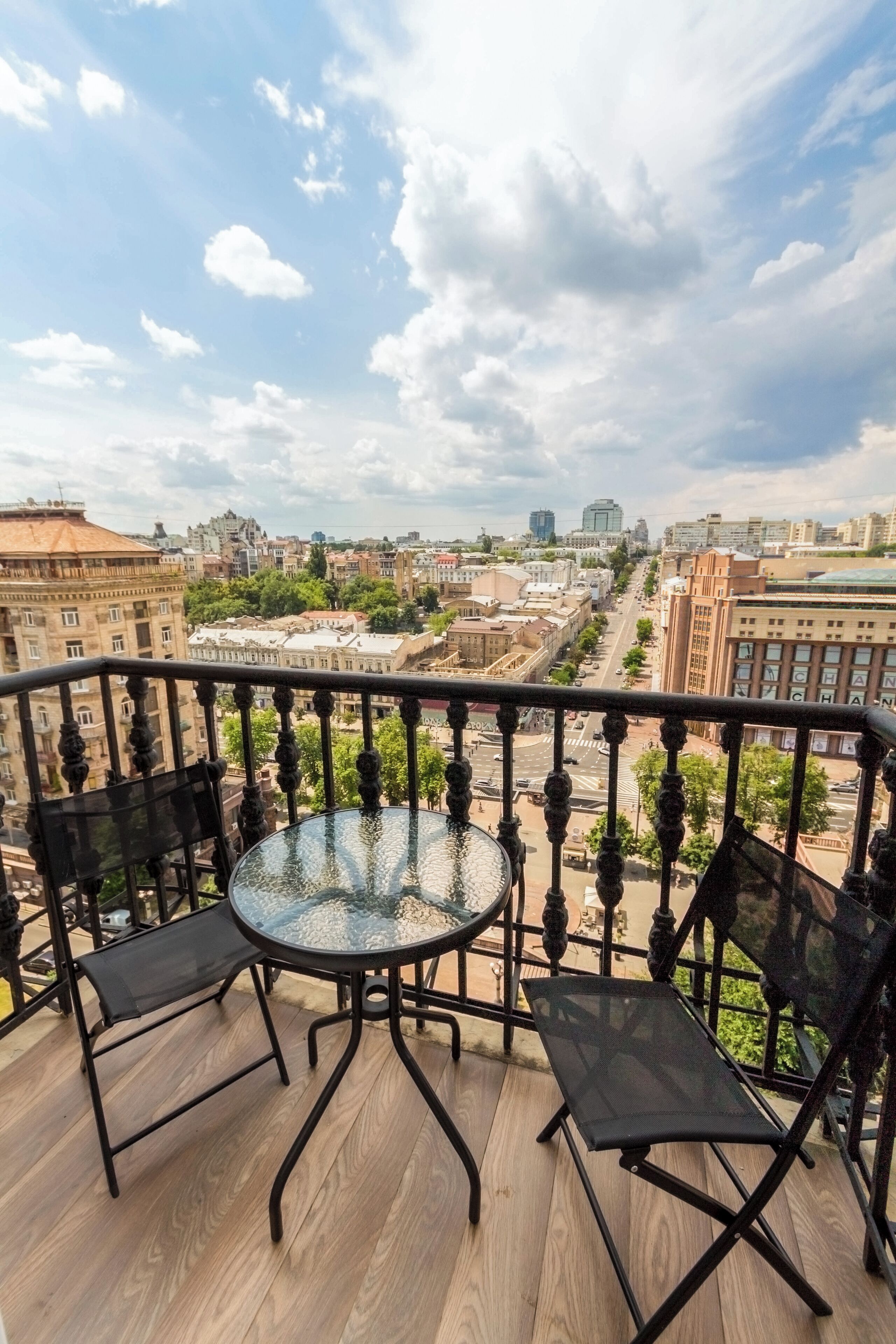 A balcony with a round glass table and two black chairs, overlooking a cityscape with buildings and a clear sky.