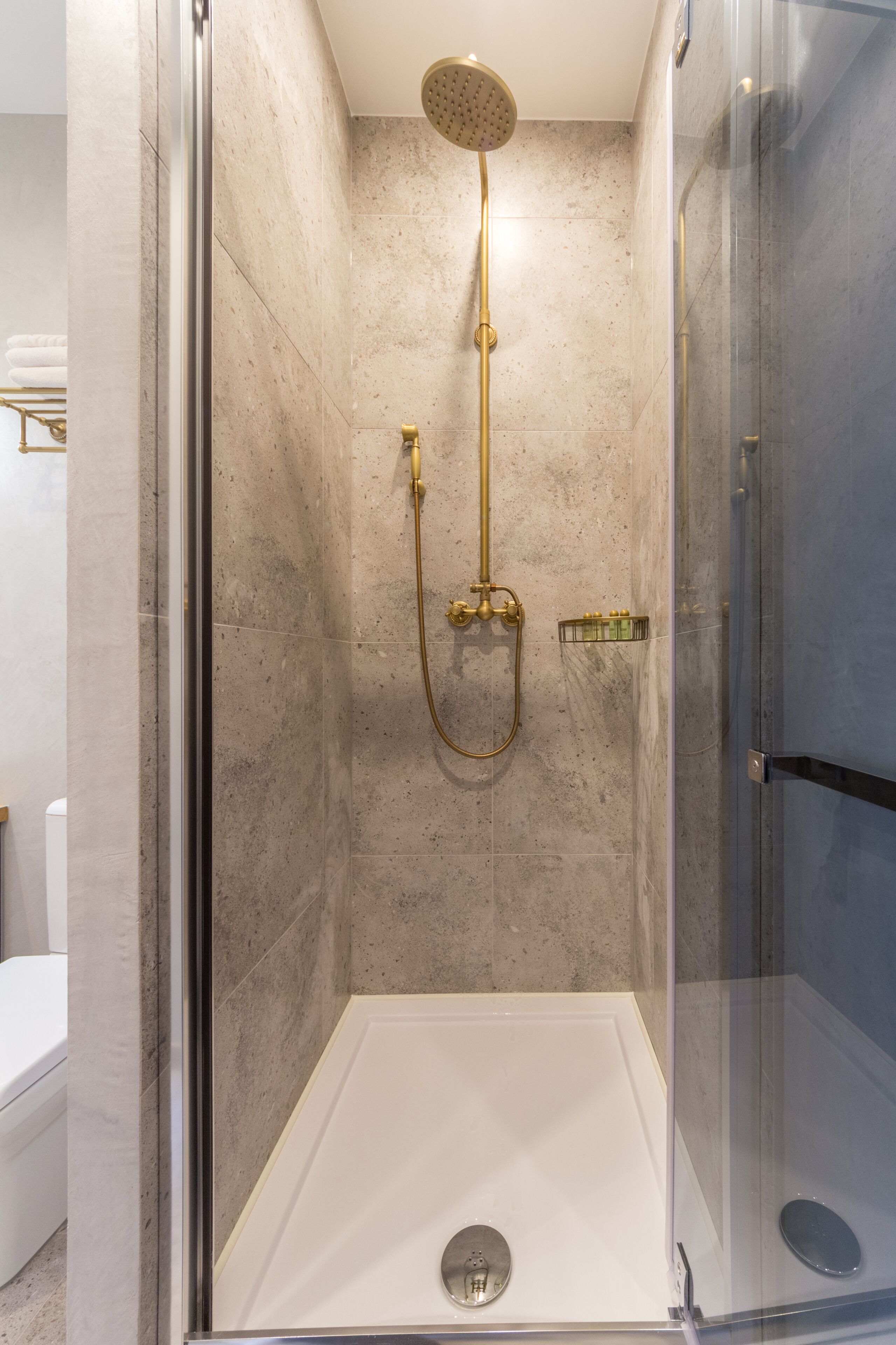 Una ducha con cerramiento de vidrio, cabezal y panel de control dorados, paredes de mármol y piso blanco.
