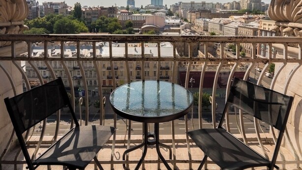 Un balcon avec une table ronde et deux chaises, donnant sur un paysage urbain avec des immeubles et des arbres.