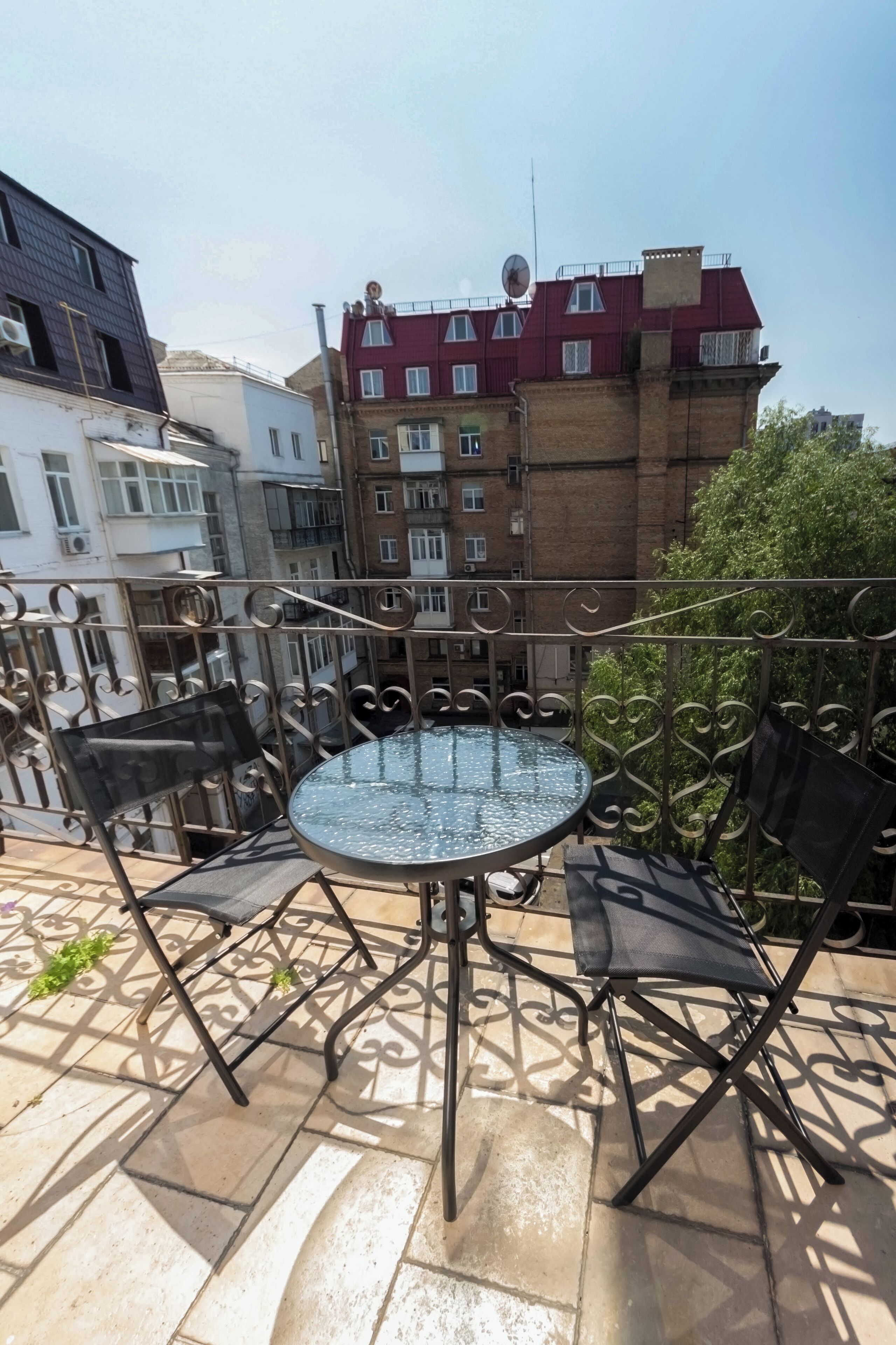 A balcony with a round glass table and four chairs, overlooking a residential area with multi-story buildings.