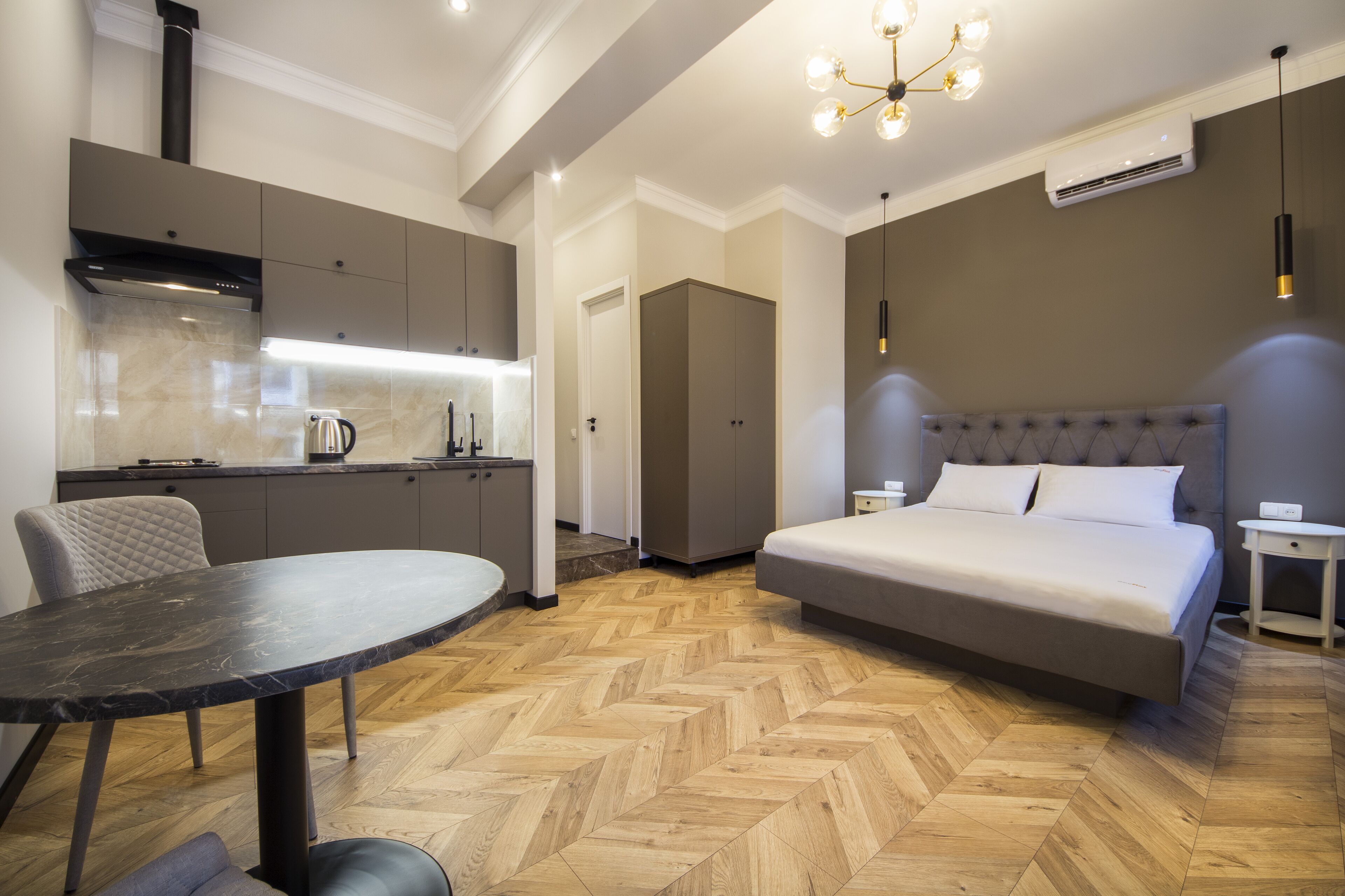 A modern hotel room with a bed, a dining table, a kitchen area, and a hallway.