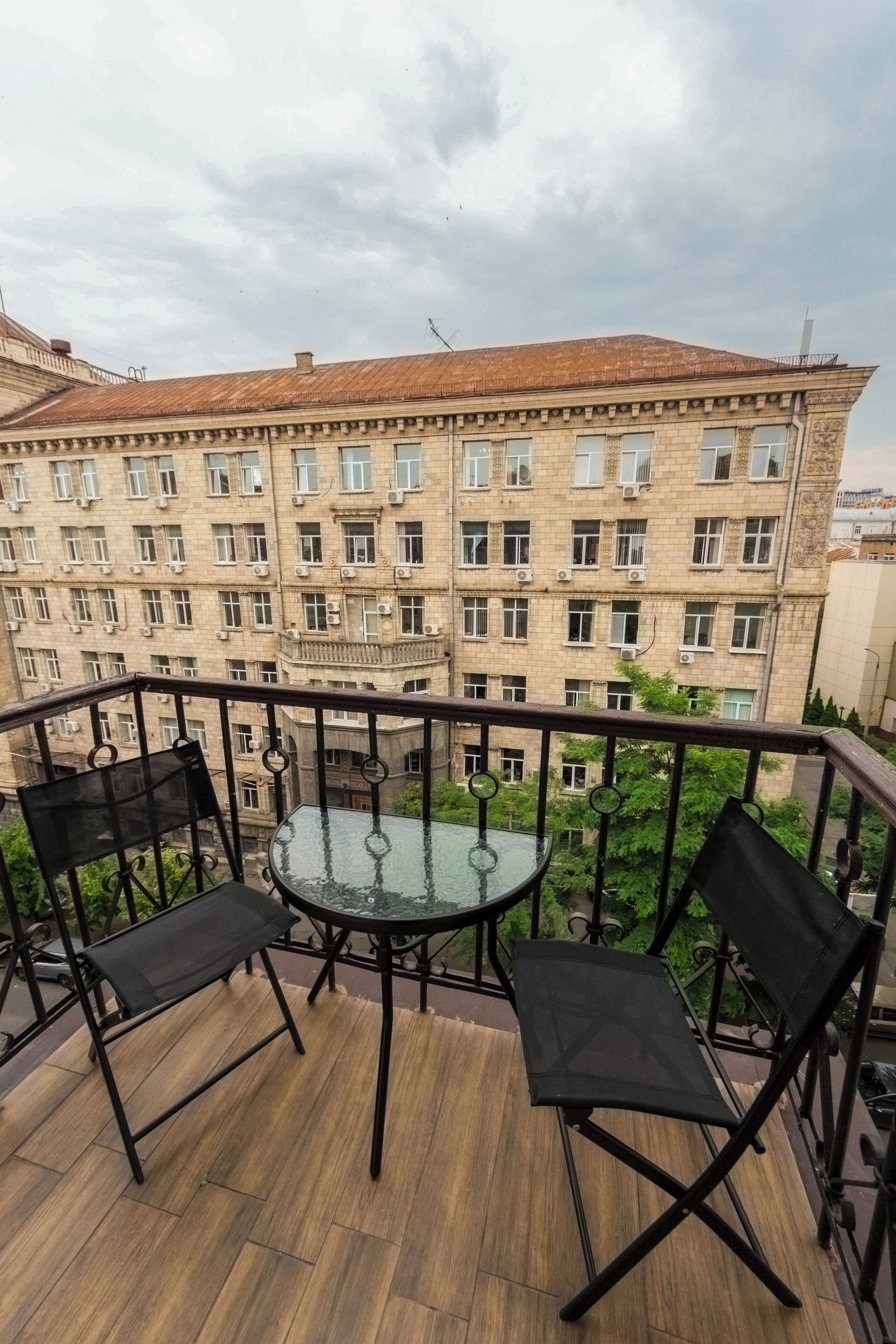 Un balcon avec une table en verre et deux chaises, donnant sur un immeuble doté de nombreuses fenêtres et de balcons.