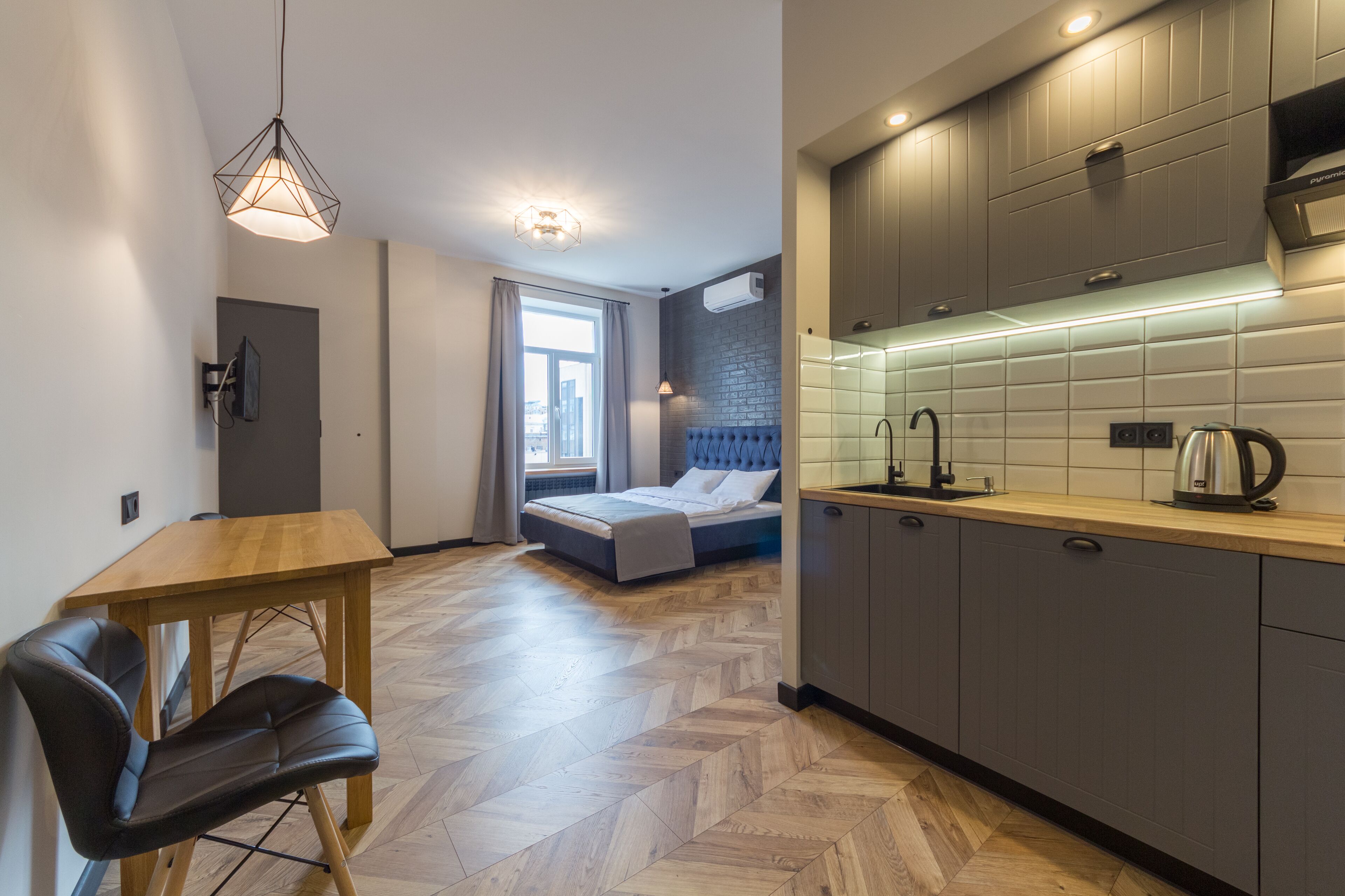 A modern kitchen with grey cabinets, a wooden countertop, and a kettle. There is a bed with a blue headboard in the background, and a dining area with a wooden table and chairs.