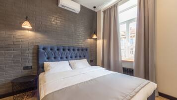 Un dormitorio moderno con pared de ladrillos, cabecera capitonada, cama grande con ropa blanca, dos lámparas colgantes y vistas a edificios a través de una ventana con cortinas.