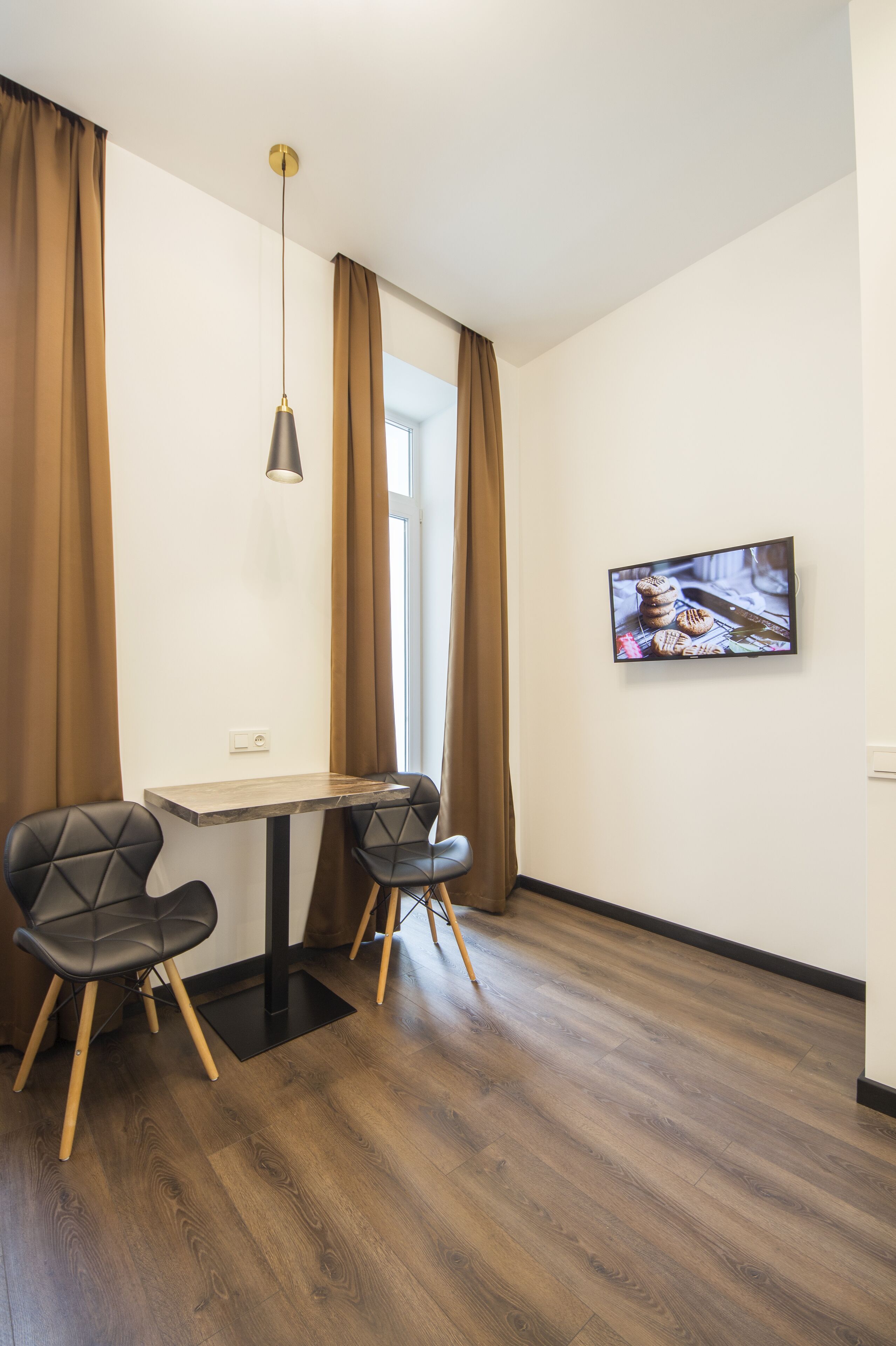 A modern room with a wooden table, two black chairs, a hanging pendant light, and a flat-screen TV on the wall.