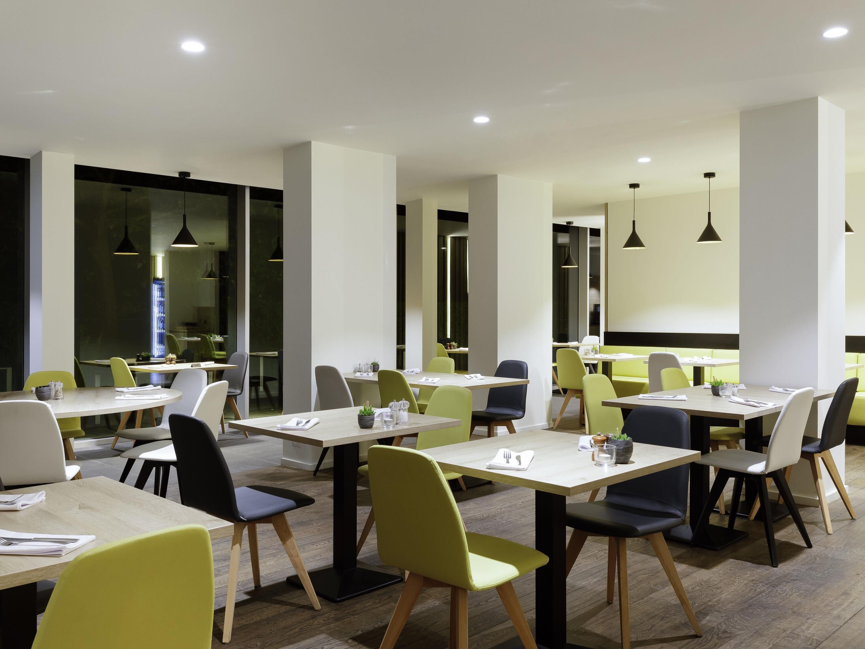 A modern dining area with multiple tables and chairs, featuring a clean, minimalist design with natural wood flooring and pendant lighting.