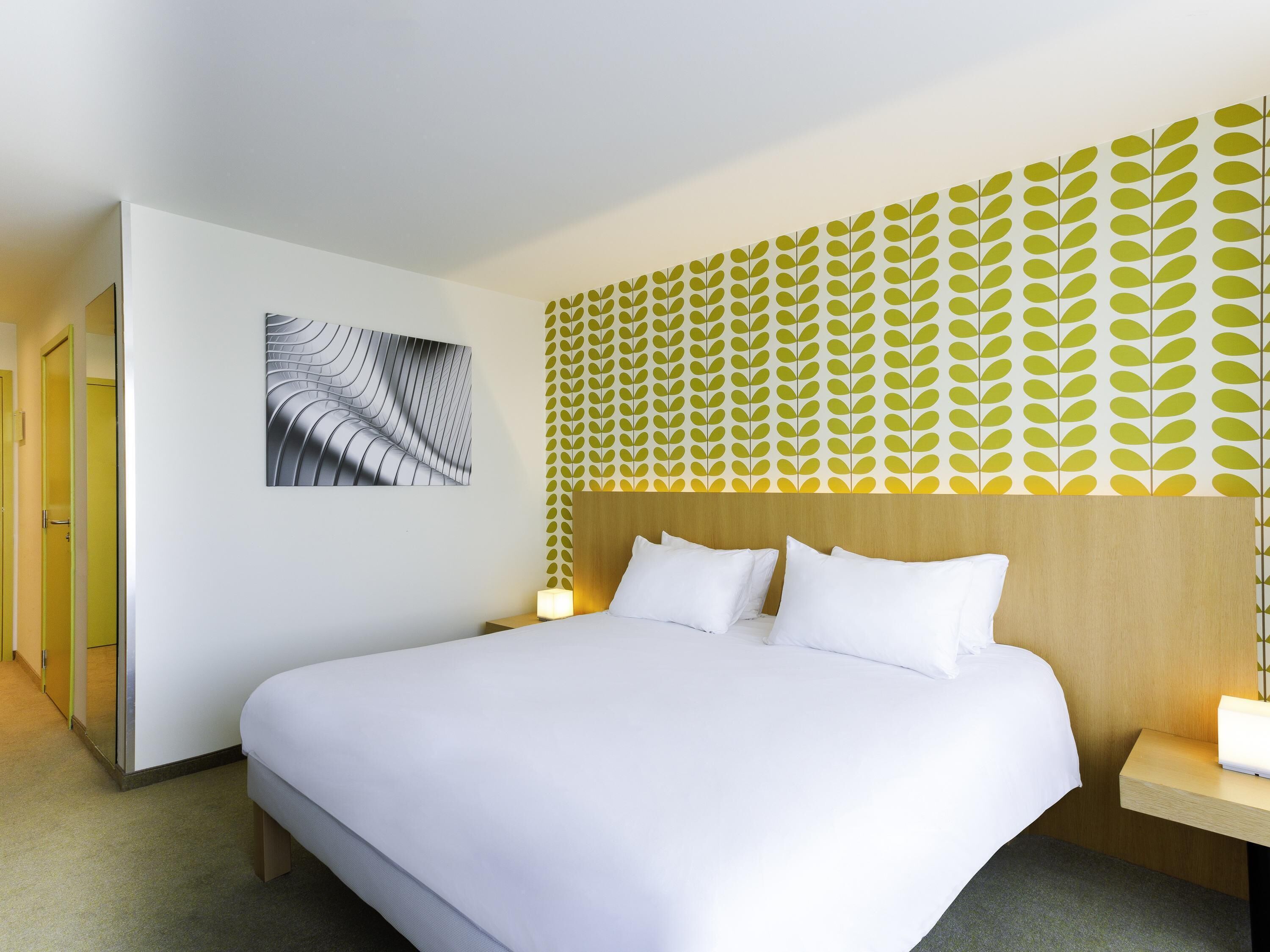 A hotel room with a large bed, a bedside table with a lamp, a wall-mounted artwork, and a patterned wallpaper design.