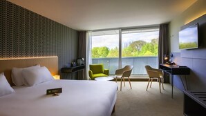 Ein Hotelzimmer mit einem großen Bett, einem Schreibtisch mit Flachbildfernseher, einer Sitzecke mit Stühlen und einem Balkon mit Blick auf Bäume.