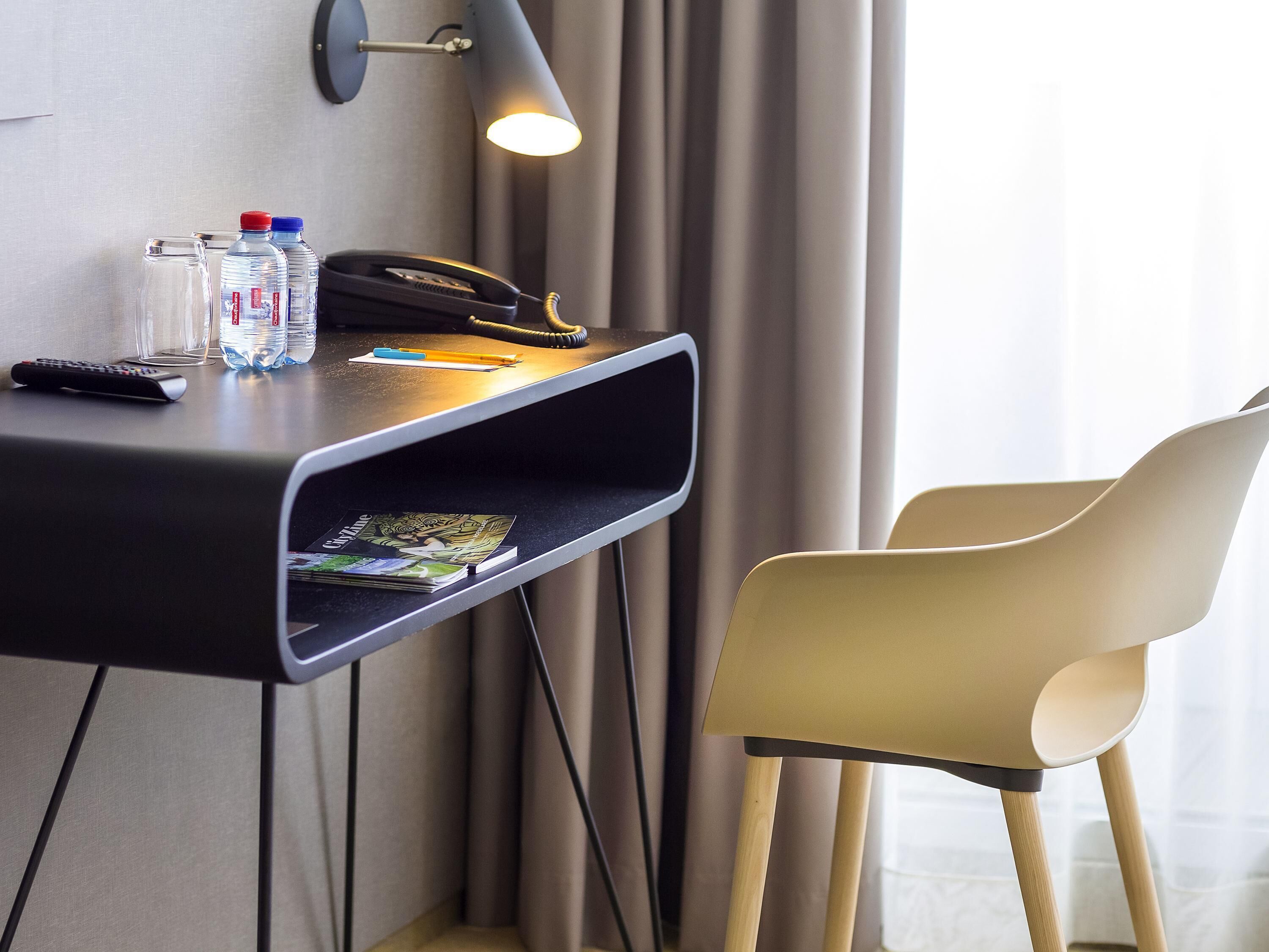 A hotel desk with a telephone, remote control, water bottles, and a magazine.