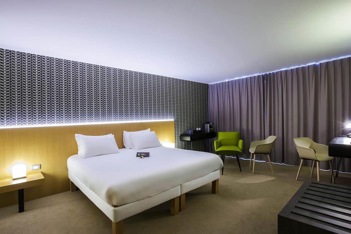 A hotel room with a bed, bedside tables, a desk, chairs, and a patterned wall.