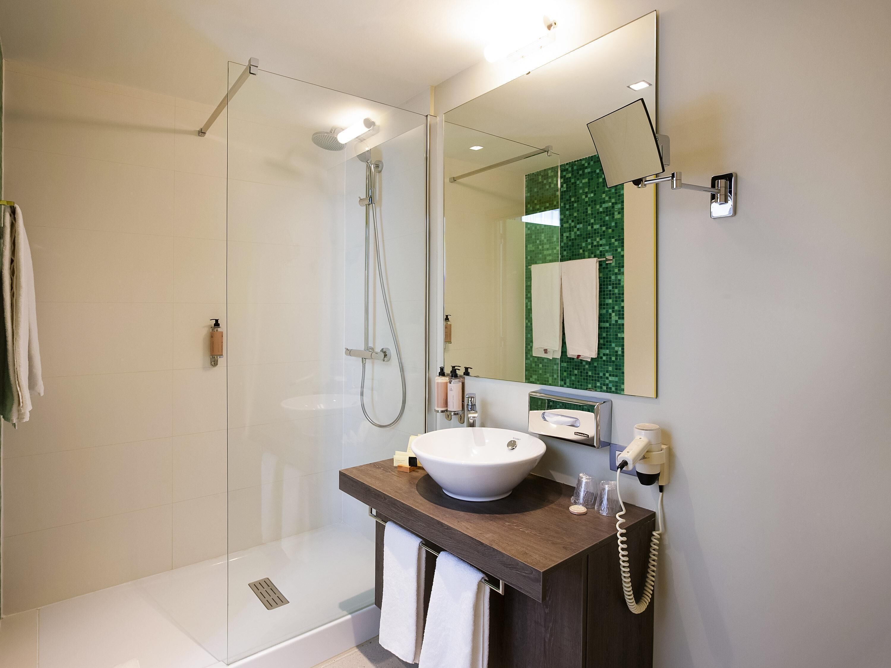Een moderne badkamer met een douchecabine van glas, een witte wastafel en een spiegel met een groene mozaïekachtergrond.