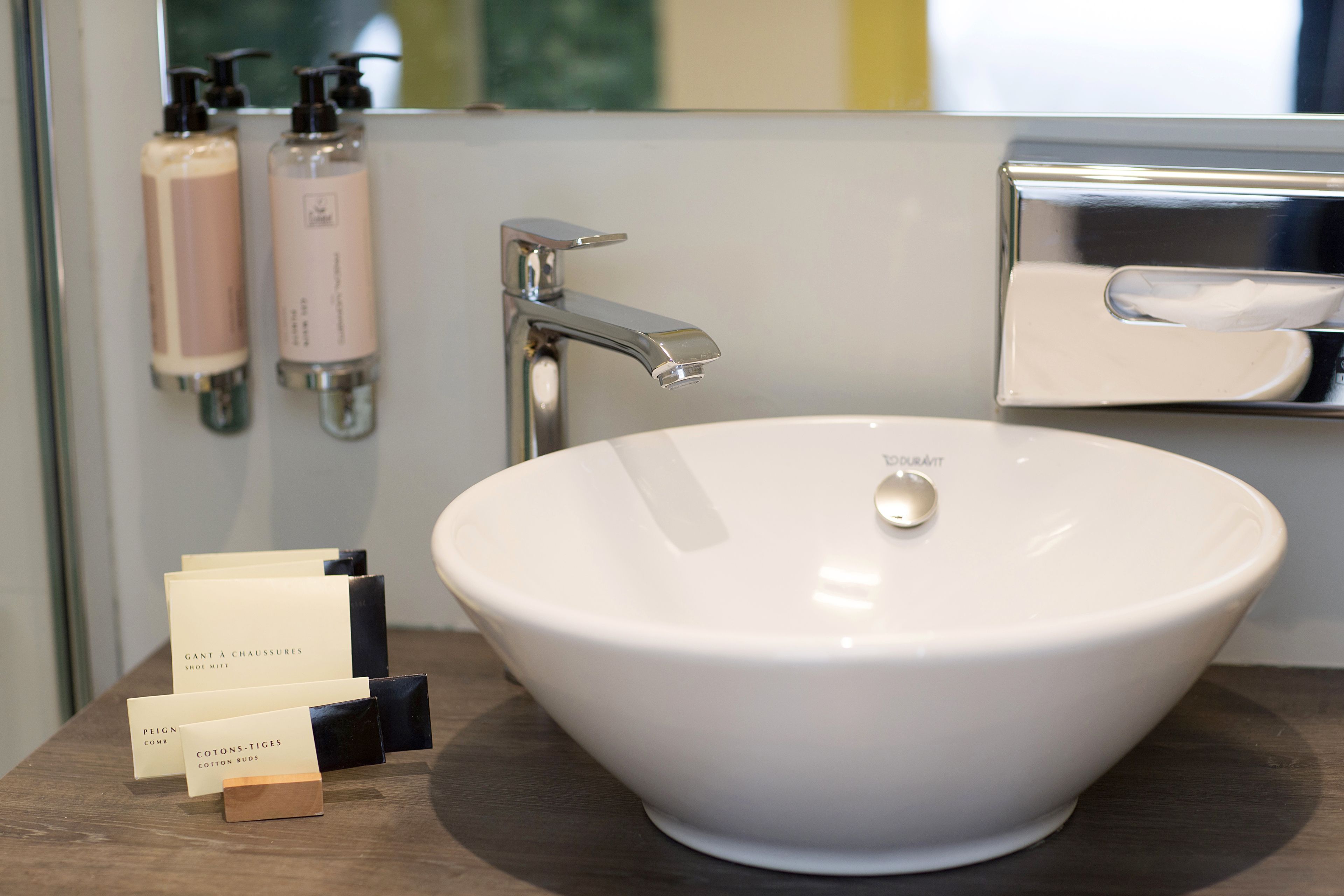 A bathroom with a white sink, a chrome faucet, and a mirror with a soap dispenser. There are also bottles of lotion and a small box of cotton buds.