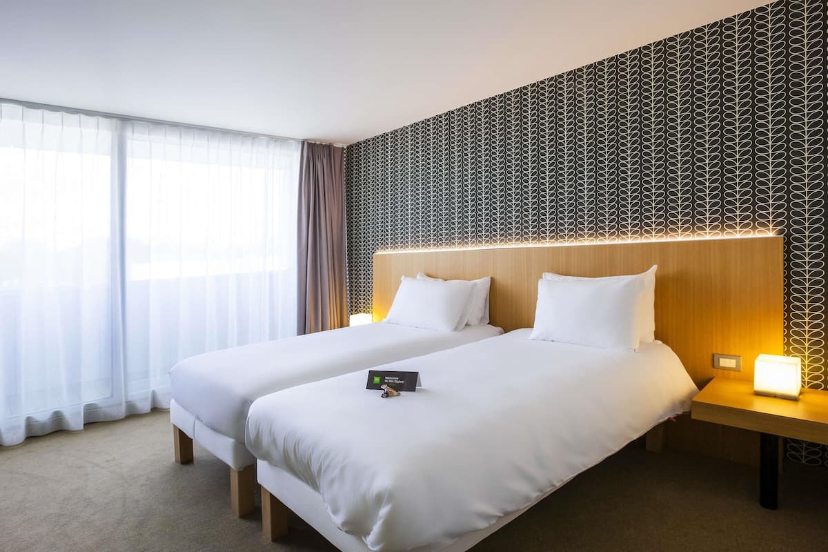 Hotellihuone, jossa on kaksi sänkyä, suuri ikkuna läpikuultavilla verhoilla, kuvioitu seinä ja yöpöytä, jossa on lamppu.
