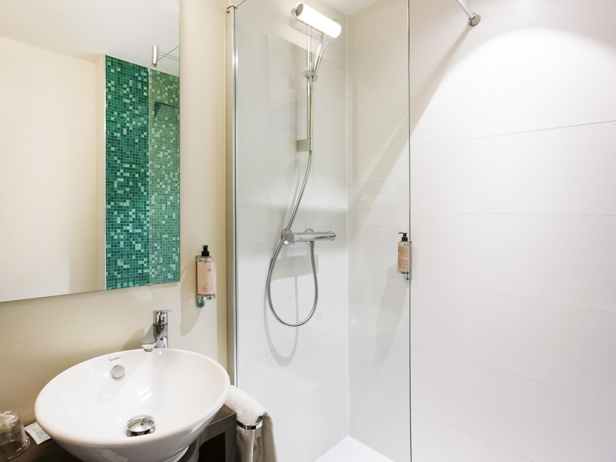 Une salle de bain avec un lavabo blanc, une cabine de douche en verre et un mur recouvert de mosaïques vertes.
